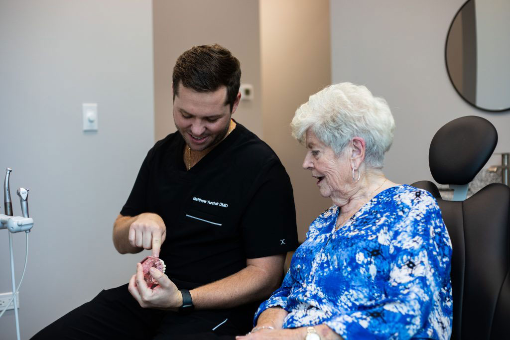 This is a photograph showing an older woman seated on a dental chair with her eyes closed, while a male dental professional stands beside her, holding up a mirror and examining her mouth.