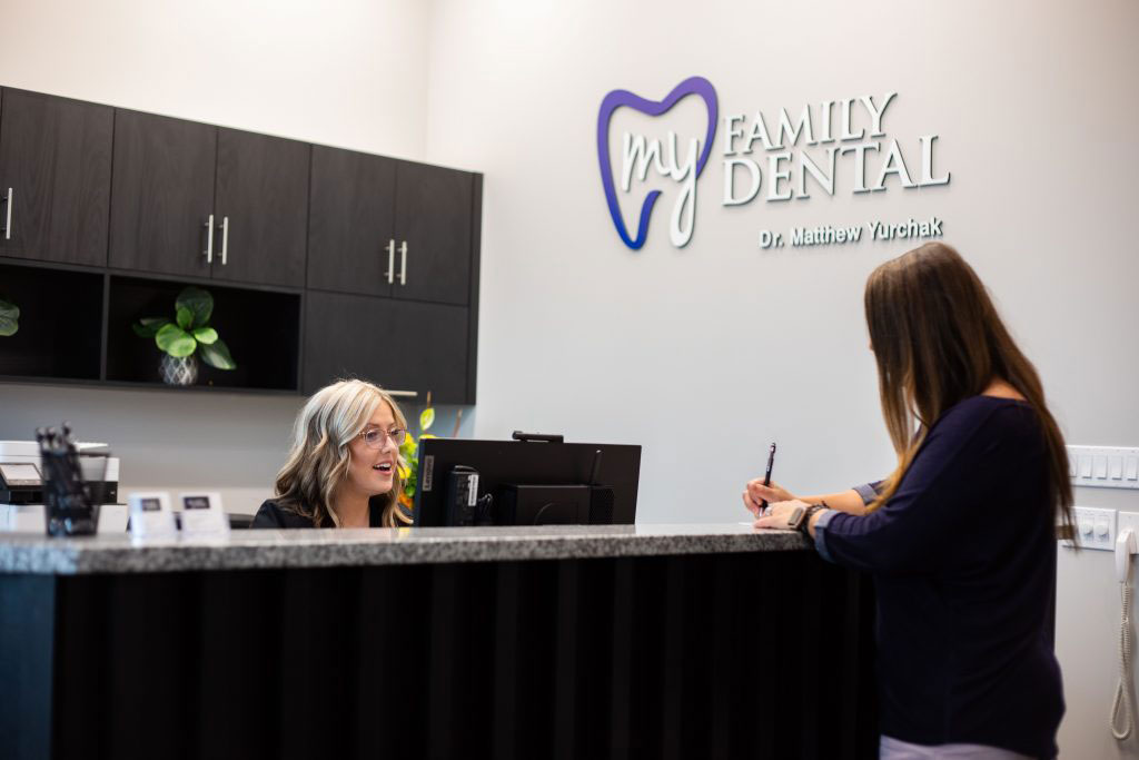 The image shows an interior view of a dental office reception area with a woman standing at the counter, a sign for  Wau Dental  on the wall behind her, and a person sitting at the desk in front of her.