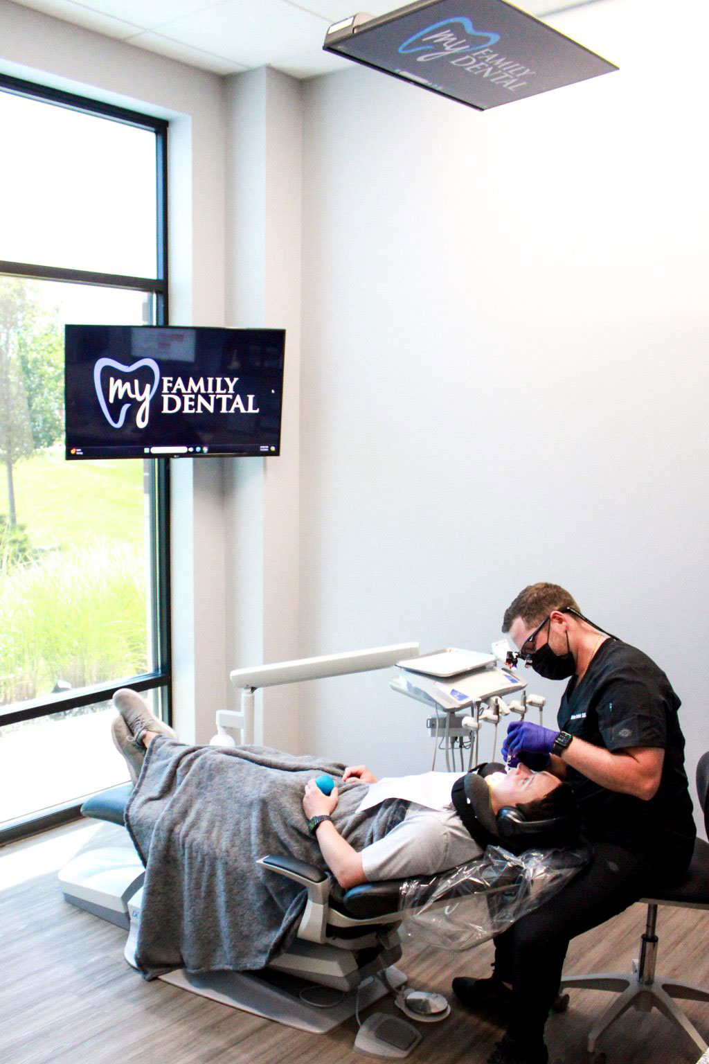 A dental office with a dentist performing a procedure on a patient while another person monitors the equipment.