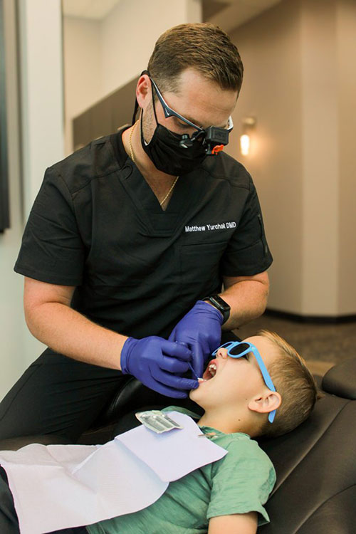 A dental professional is performing oral care on a young child while wearing protective gear.