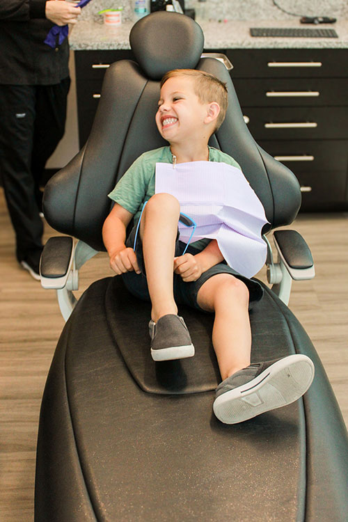 A young boy sitting on a dental chair with a paper draped over his face, smiling at the camera.