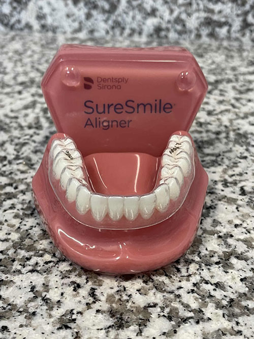 The image shows a dental appliance, specifically an upper denture, with a set of artificial teeth and gums, placed inside a pink case with text indicating  SureSmile Aligner.