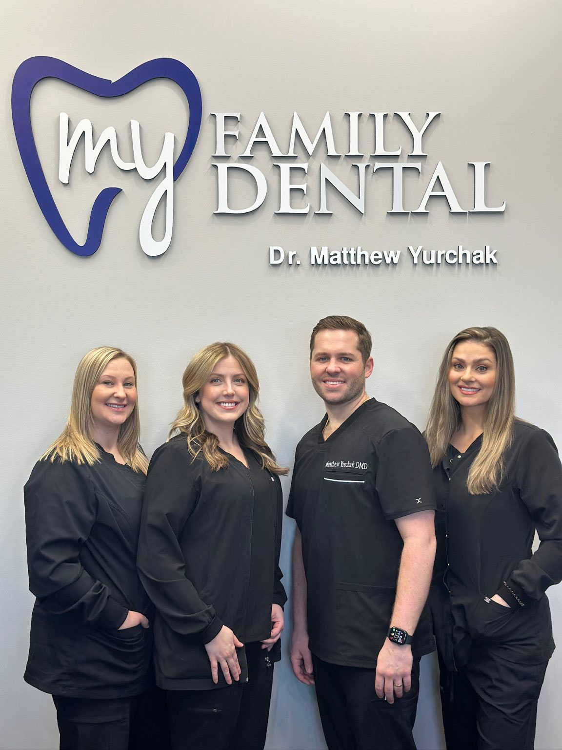 Group of four dental professionals posing together in front of a logo for  My Family Dental  with a tagline.