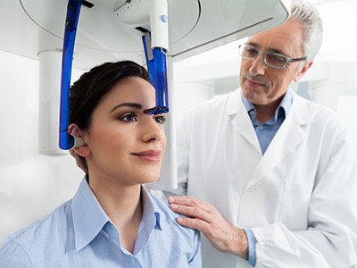 An image of a woman seated in a dental chair with a device attached to her head, receiving care from a dentist wearing a white coat and stethoscope.