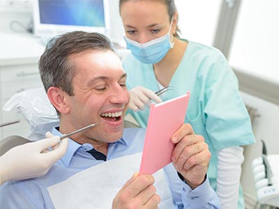 A man sitting in a dentist s chair, holding up a pink card with a surprised expression, while a dental professional looks on with a stethoscope around their neck.