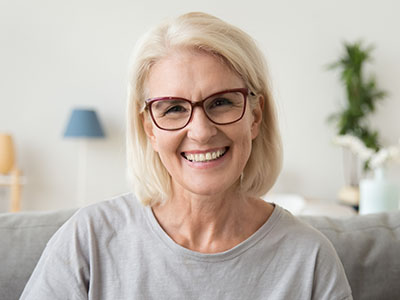 The image shows a smiling woman with short hair wearing glasses, sitting on a couch indoors with a warm ambiance.