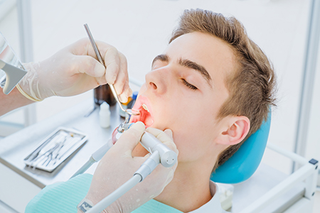 The image shows a person receiving dental care while seated in a dental chair, with a dental professional using a drill on their tooth, surrounded by dental equipment and tools.