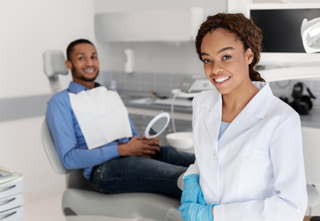 The image shows a dental office setting with a woman wearing a white lab coat and a smiling man in a blue shirt sitting in a dental chair, both looking at the camera.