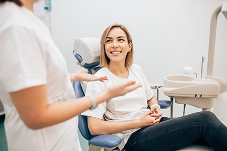 The image shows a dental office setting with a woman seated in a dental chair, smiling at the camera, while a dental hygienist stands beside her, gesturing towards her mouth with one hand.
