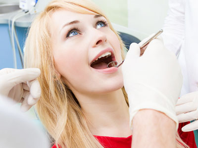 The image shows a woman undergoing dental treatment, with her mouth open wide, revealing a dental drill being used on her teeth by a dentist wearing gloves and a face mask.