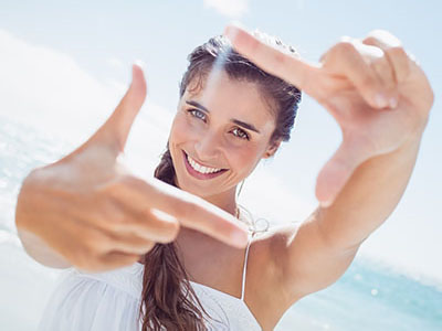 The image shows a smiling woman with long hair, taking a selfie with her right hand while holding up a peace sign with her left hand. She is outdoors, wearing a white top, and appears to be on a sunny day at a beach setting.